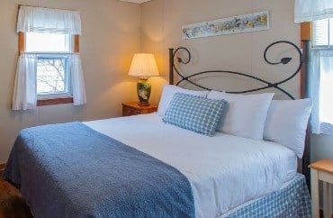 Small bedroom with queen bed in white and blue linens, side table with lamp and windows with white curtains